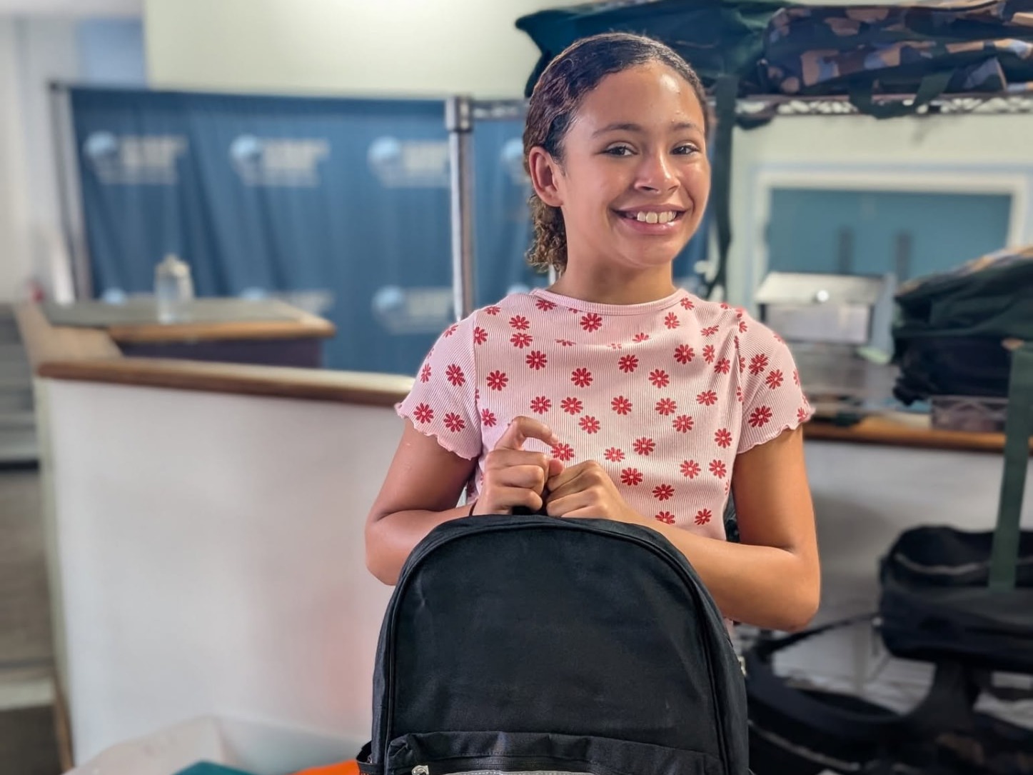 A smiling student holding a new black backpack at the Connect Center, with bins of supplies behind her